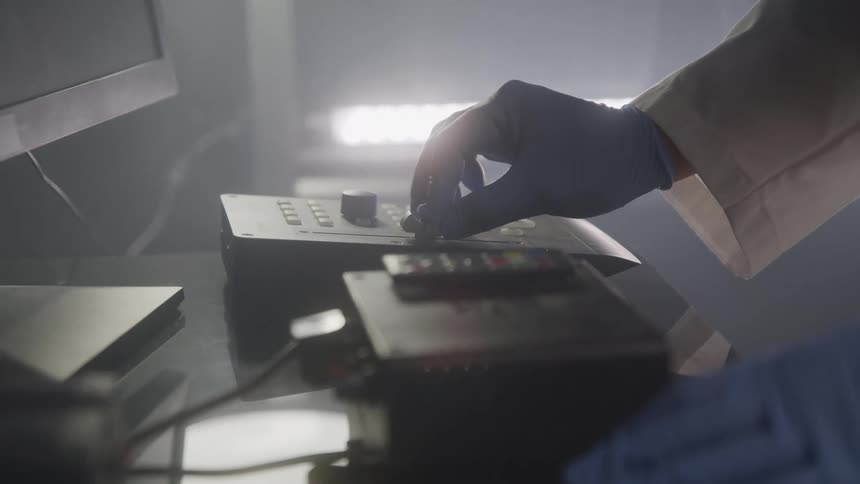 A closeup shot shows a gloved hand operating a control panel with various buttons and a joysticklike control the environment appears to be dimly lit with some bright lights in the background
