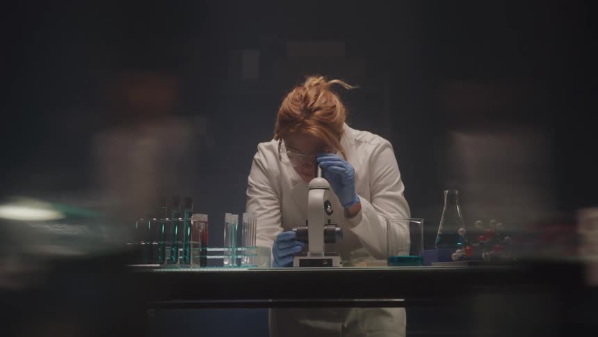 A female scientist wearing a white lab coat safety glasses and blue gloves is shown intently looking through a microscope she is surrounded by various laboratory equipment including test tubes and beakers with colored liquids