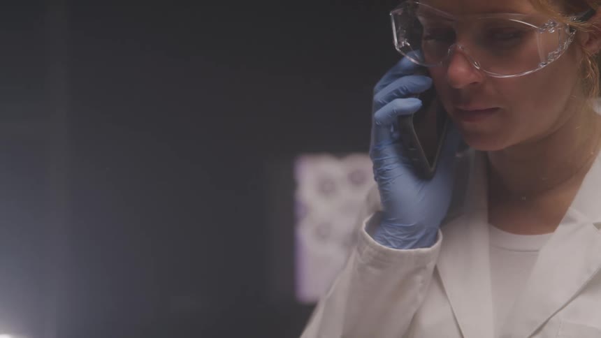 The video features a woman in a lab coat and safety goggles holding a phone to her ear with a gloved hand suggesting she is in a scientific or medical setting she appears focused and engaged in a serious conversation