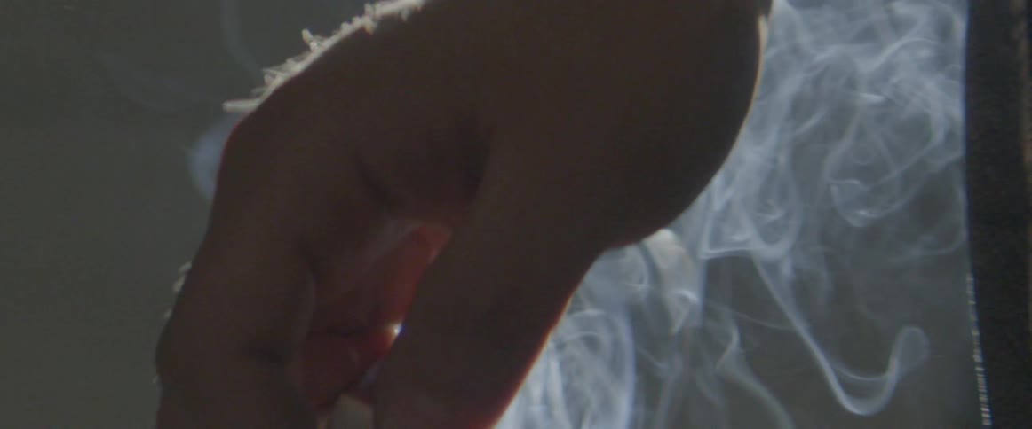 A hand holds a burning cigarette as smoke swirls around