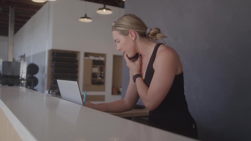 A woman with blonde hair tied back is speaking on a phone while also looking at a laptop she is standing at a counter in what appears to be a modern or industrialstyle space