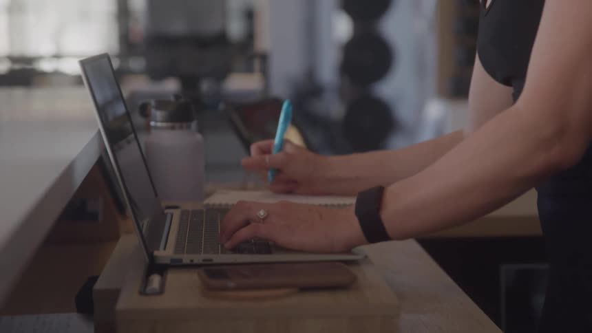 A woman with a ponytail wearing a black tank top and a smartwatch types on a laptop and takes notes with a blue pen at a wooden counter