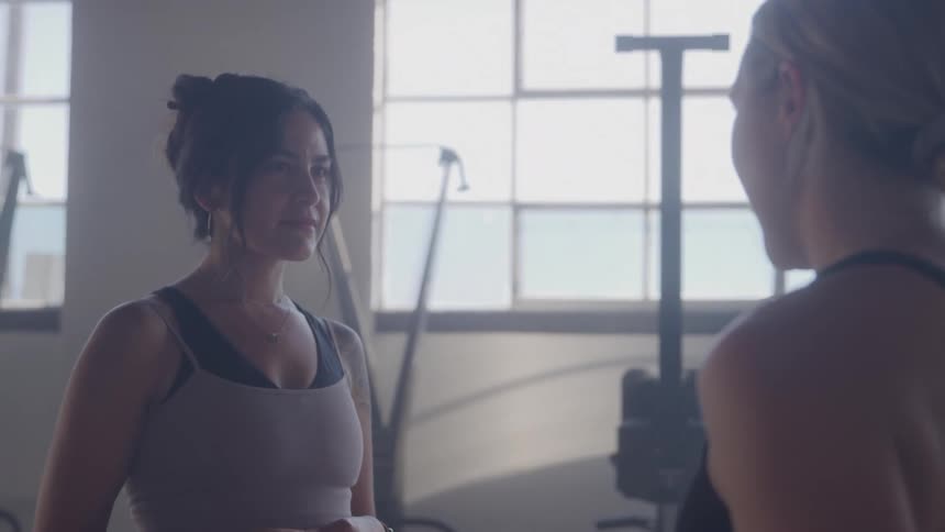 Two women talk in a gym lit by natural light from the windows