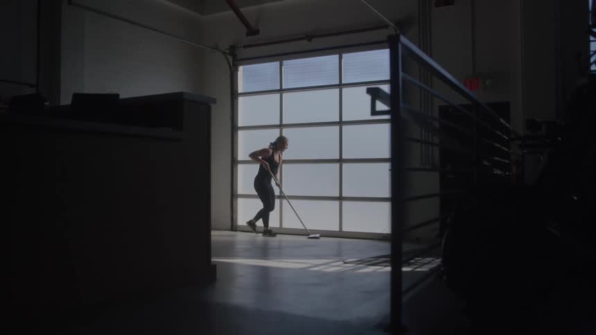 A person mops the floor in a dimly lit industrial space with sunlight streaming through large frosted windows