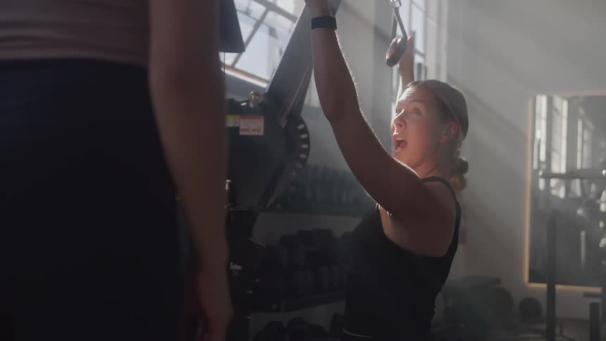 A young woman demonstrates how to use a gym equipment to another woman in the gym during daytime