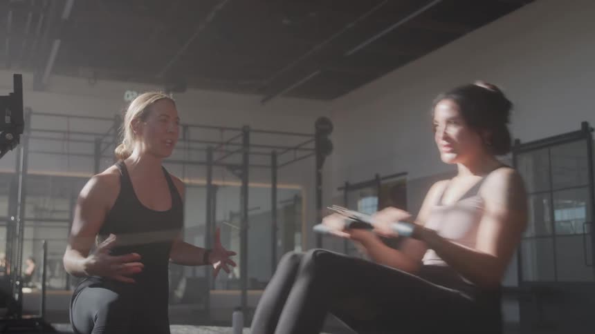 A woman exercises on a rowing machine while another woman coaches her