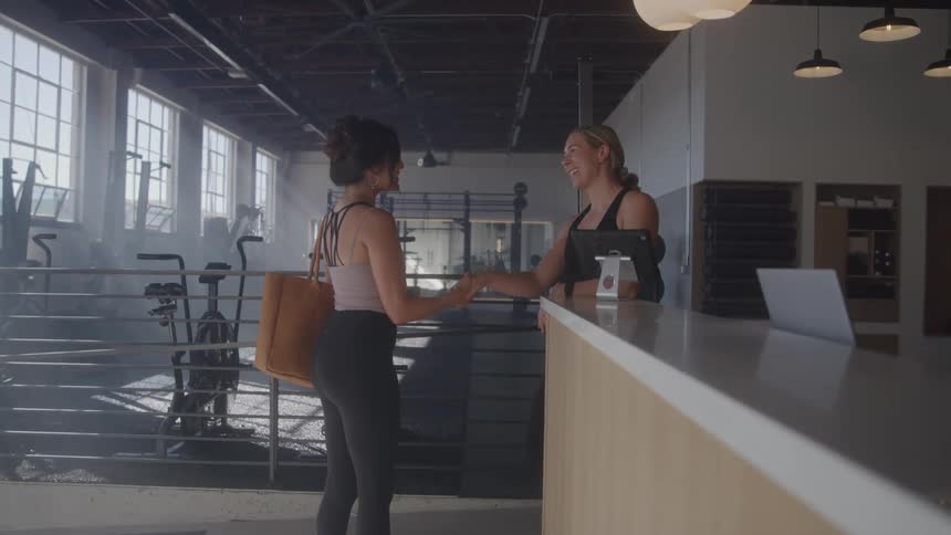 The video shows two women interacting at a reception desk within a spacious gym or fitness studio one woman presumably a gymgoer approaches the desk and engages in a friendly conversation with the other woman who appears to be a staff member
