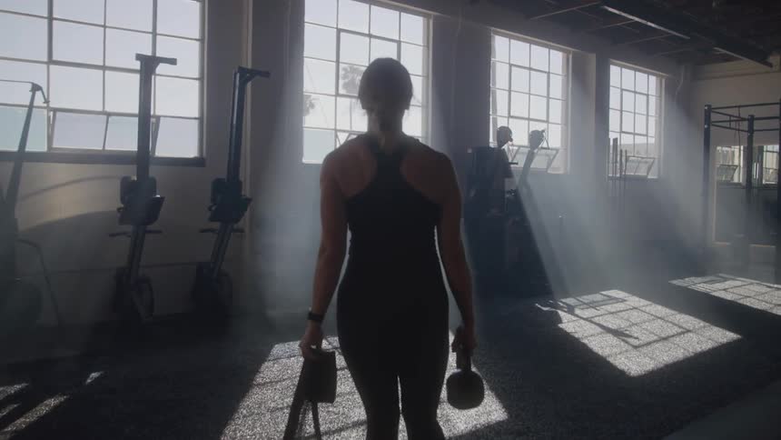 A woman in workout attire walks towards large sunlit windows in a gym carrying a resistance band and a kettlebell as dust motes dance in the light rays