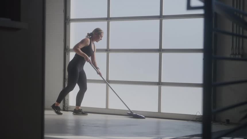 A woman in dark workout clothes is shown mopping a large floor area with a wide mop behind her a large window provides ample natural light to the space