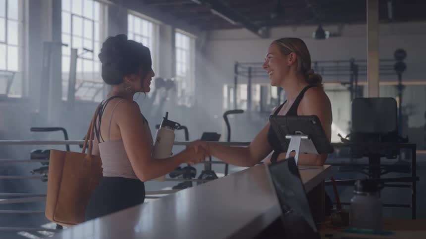 Two women smile and chat while shaking hands at a gym front desk
