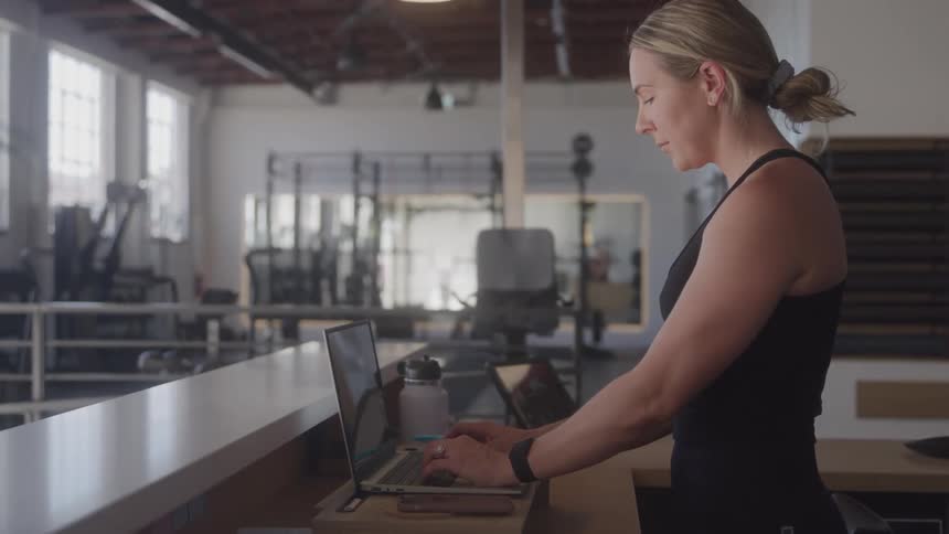 A woman works intently on a laptop at a counter in a gym setting