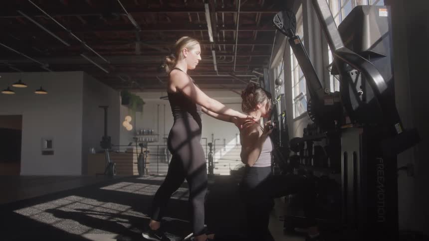 The video shows two women in a gym setting one woman wearing black workout attire appears to be a trainer guiding or assisting another woman who is using a cable machine for an exercise sunlight streams in from windows casting shadows on the floor