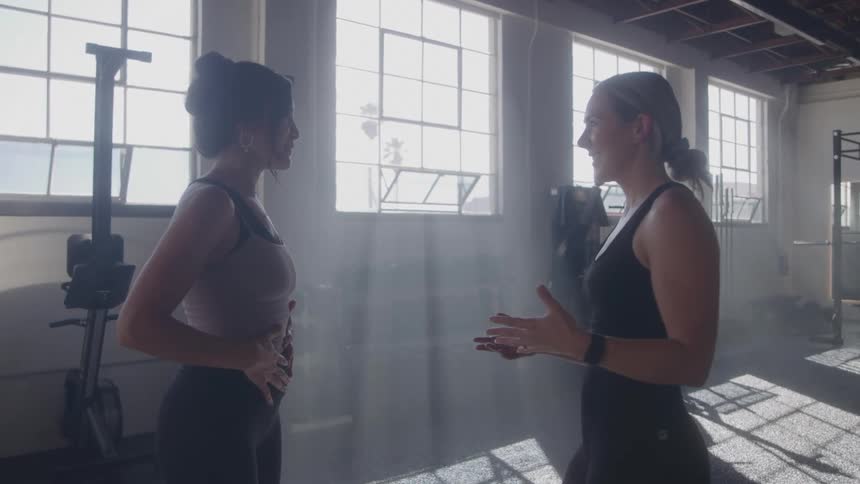 Two young women talk to each other in a gym at midday as they prepare for a workout