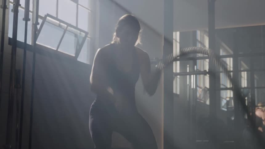 The video features a woman in a gym setting vigorously exercising with battle ropes the strong backlighting from a large window creates a striking silhouette emphasizing her movements