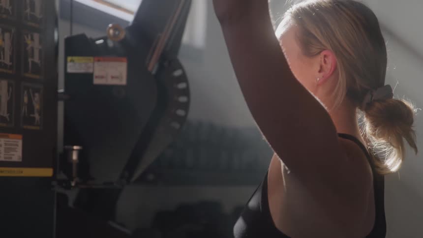 The video shows a woman with blonde hair tied back wearing a black sports bra and a fitness tracker exercising on a cable machine at a gym she is seen from the side with her muscles visibly engaged as she performs repetitions