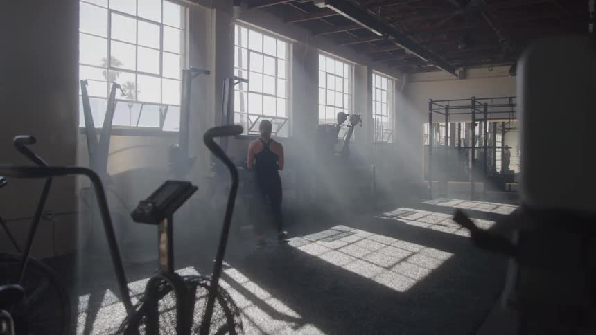 Gym employee looks over the equipment in a gym while the sunlight entering through nearby windows creates shadows on the floor