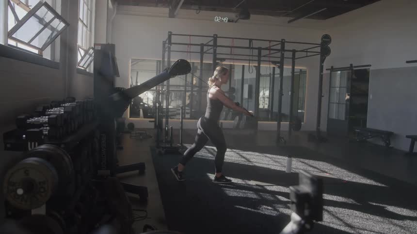 A blonde woman is exercising in a gym pulling on a resistance band attached to a weight rack the gym features exposed ceiling beams and large windows with various exercise equipment visible in the background