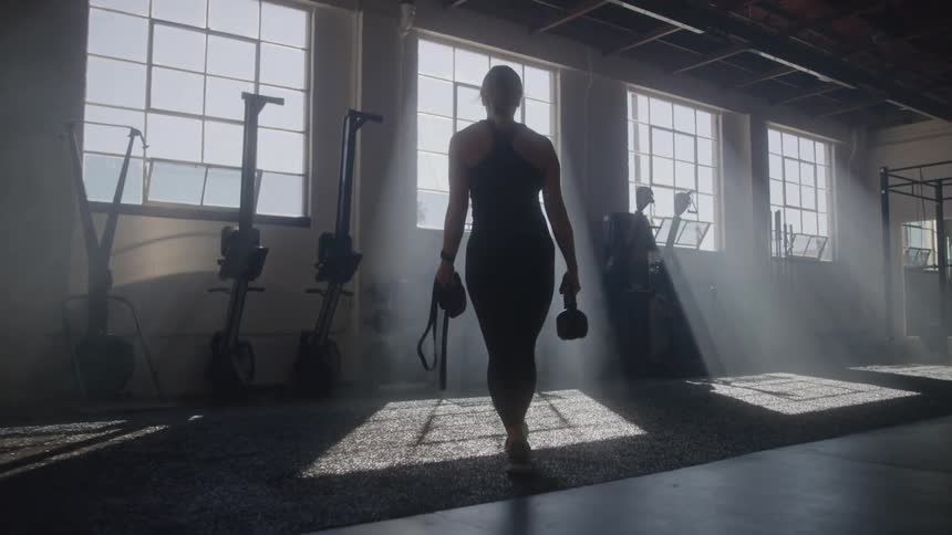 The woman walks towards the weights in the gym