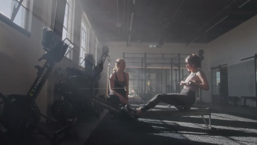 Two women in a sunlit gym one using a rowing machine while the other observes and coaches