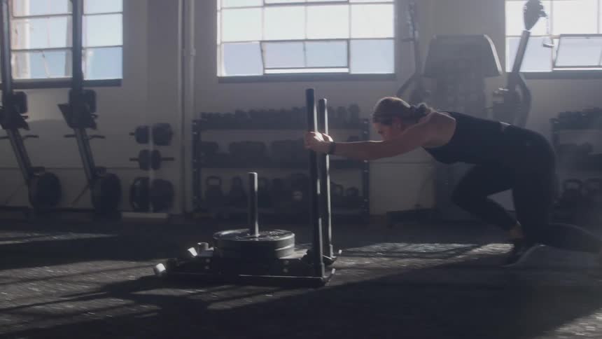 A woman in black activewear is seen pushing a weighted sled across a gym floor sunlight streams in through large windows in the background illuminating the dusty air and gym equipment