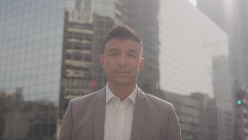 Man in beige blazer in an outdoor setting with buildings in the background
