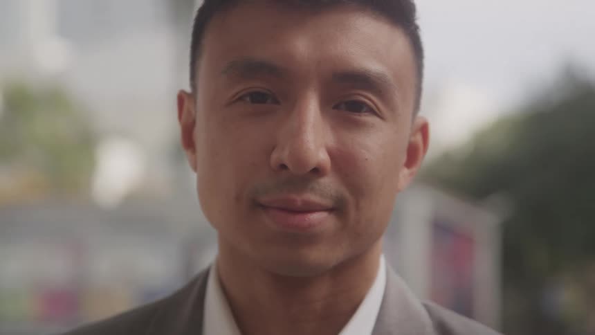 A man in a suit looks at the camera with a calm expression in an outdoor urban setting