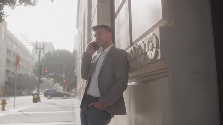 A man in a blazer and jeans is leaning against a stone building on a city street talking on his phone the background shows a sunny urban environment with trees cars and other buildings