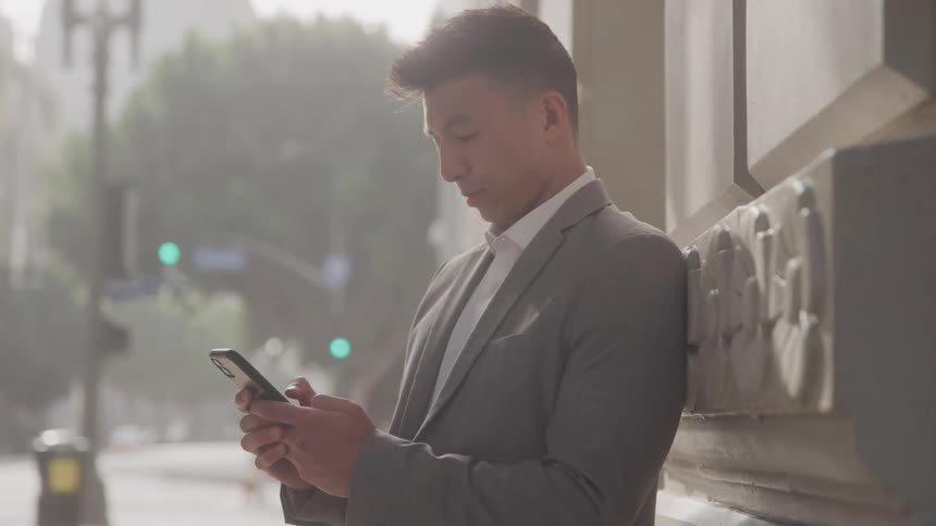Man uses both hands to tap on his phone while leaning against a building in the city