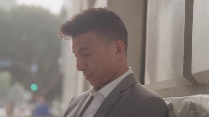 Man in a suit leans against an outdoor wall gazing thoughtfully downward