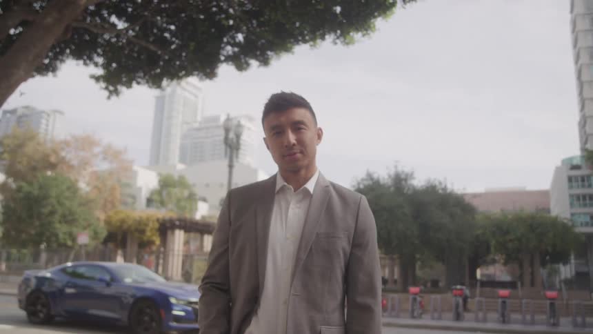 A man in a suit stands outdoors on a city street under a large tree