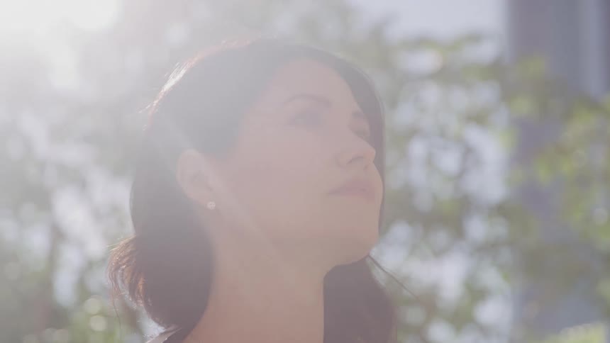 A woman with dark hair and a light colored top looks upward with a serene expression bathed in bright ethereal sunlight filtering through trees