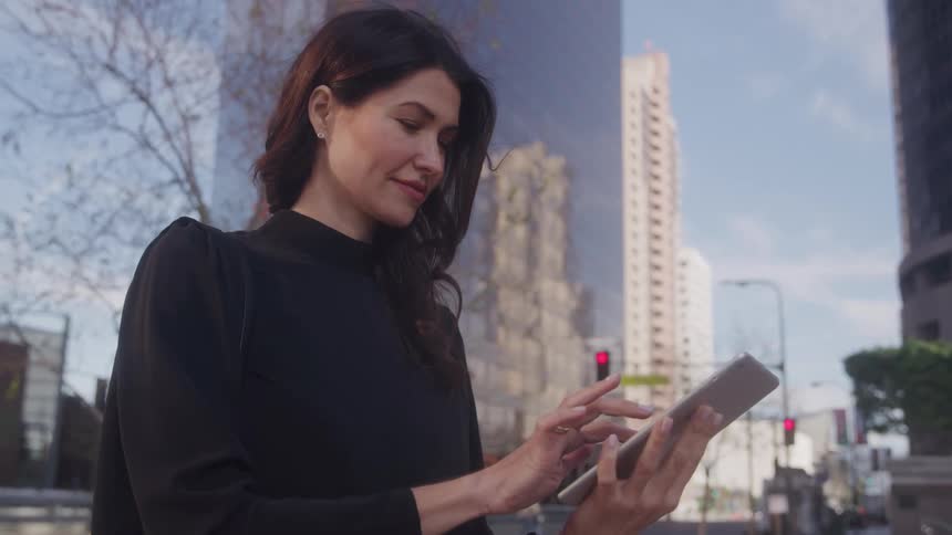 Woman sits outdoors in urban setting smiling while using tablet