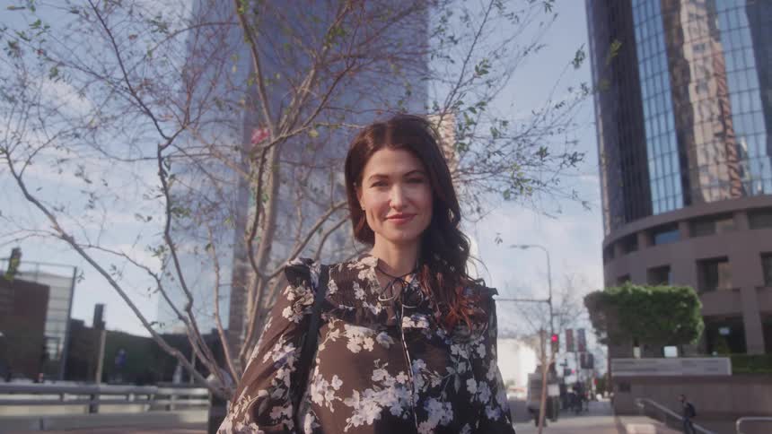 A woman with dark hair and a patterned long sleeved top stands smiling in an urban setting with tall glass buildings and a tree with bare branches in the background