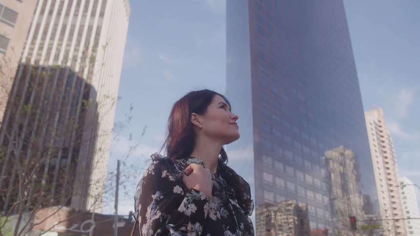 A woman in a floral blouse looks up at tall city buildings in the sunlight