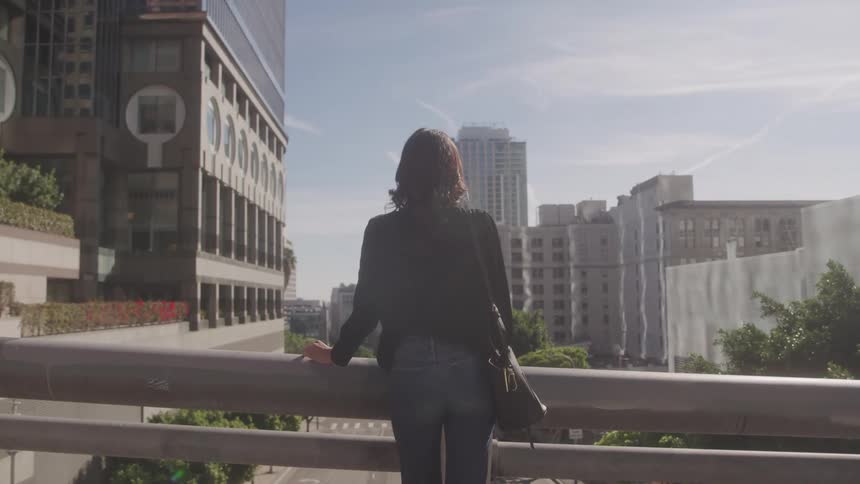 A young woman stands on a bridge holding the railing at midday in the city