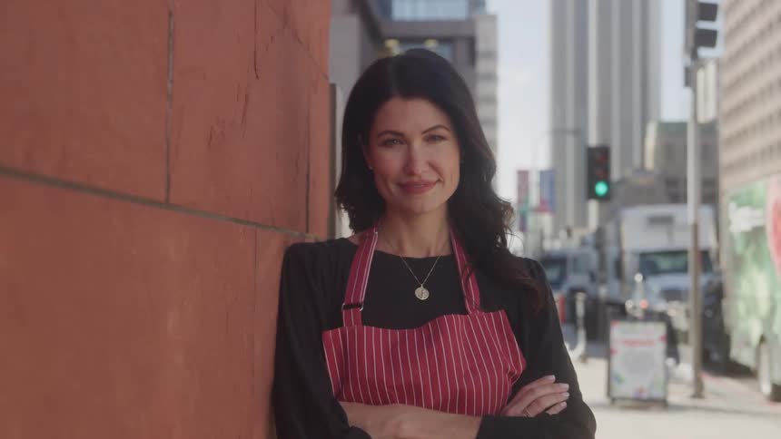 A woman wearing a necklace and apron leans against the side of a building on the street at midday