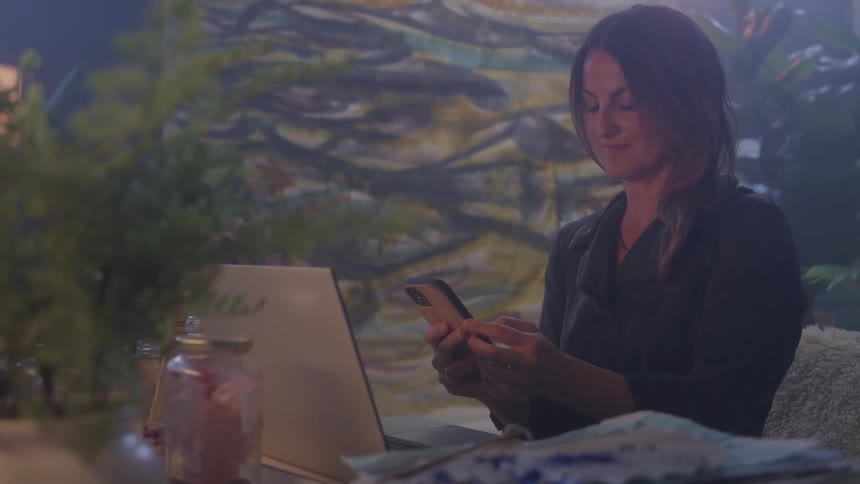 A woman sits calmly in a cozy artfilled space smiling at her phone beside an open laptop