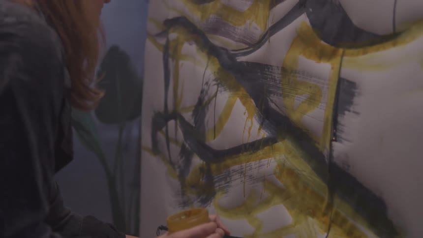 A female paints bold strokes of yellow and black on a large abstract canvas