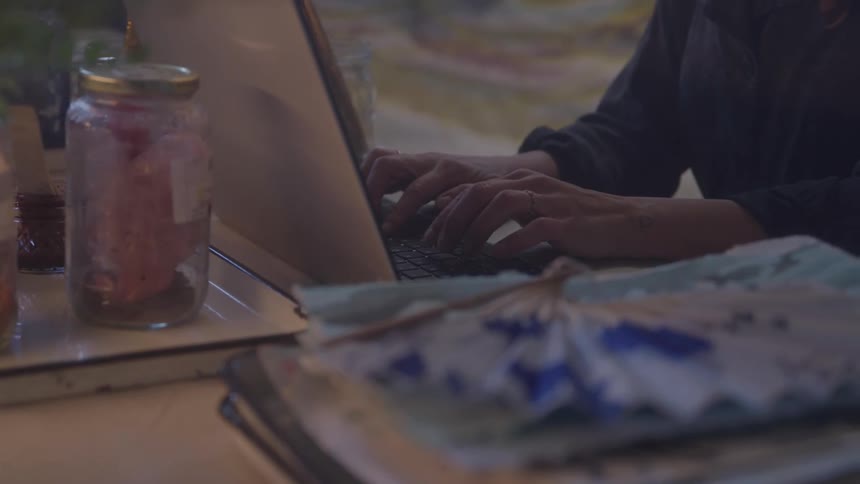 A person types on a laptop at a cluttered table with a paper fan stacked books and a jar beside them