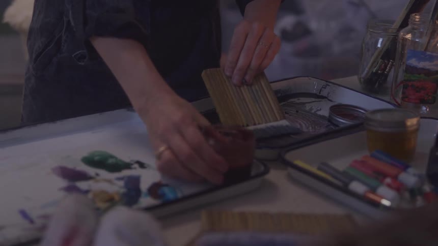 A person prepares art materials pressing a textured block into dark paint surrounded by brushes pastels and colorful supplies