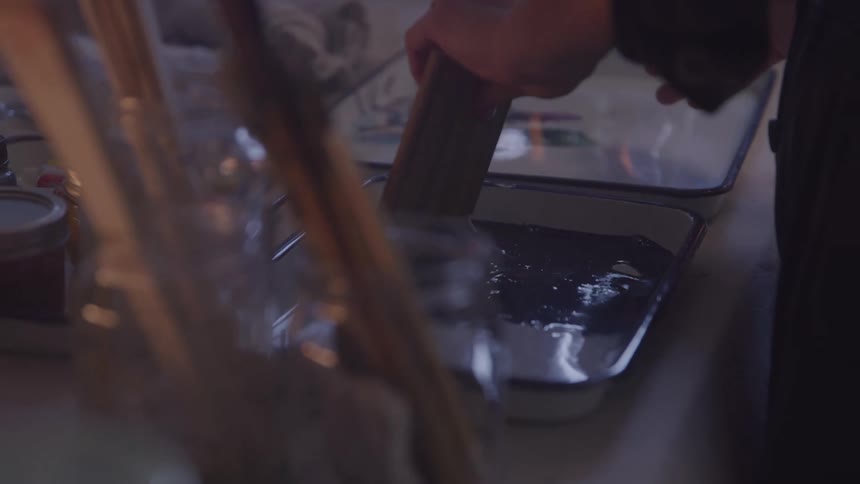 The video shows a closeup of hands pouring a dark liquid into a shallow pan possibly involving a cooking or artistic process the focus is on the action and the liquids movement with the background blurred