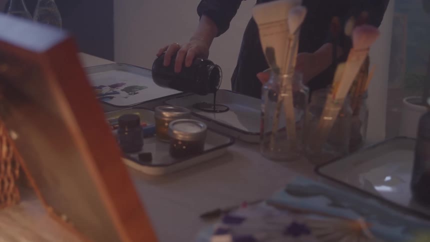 The video shows a persons hands pouring a dark liquid possibly paint or ink from a black container into a smaller jar the setting appears to be an art studio or workspace with various art supplies like paintbrushes visible in glass jars