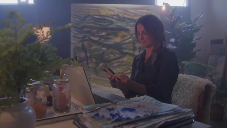 Woman sitting at a table in front of a laptop and holding a smartphone