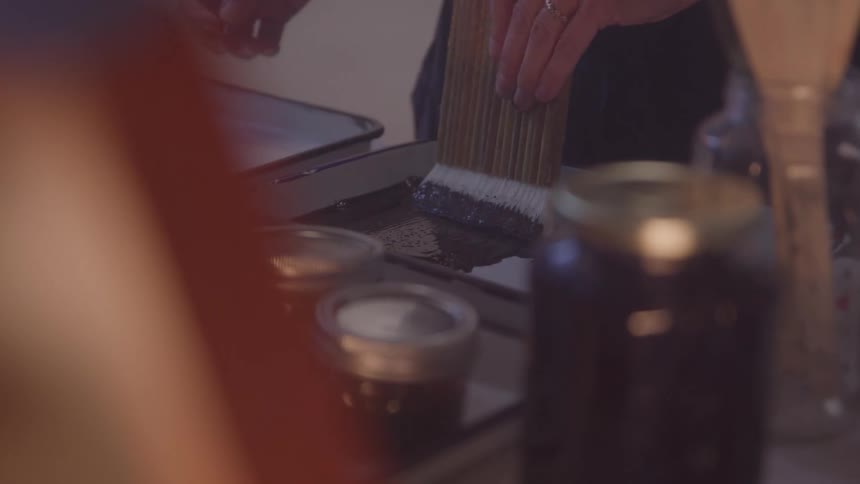 A hand brushes black ink onto a tray beside jars and art tools
