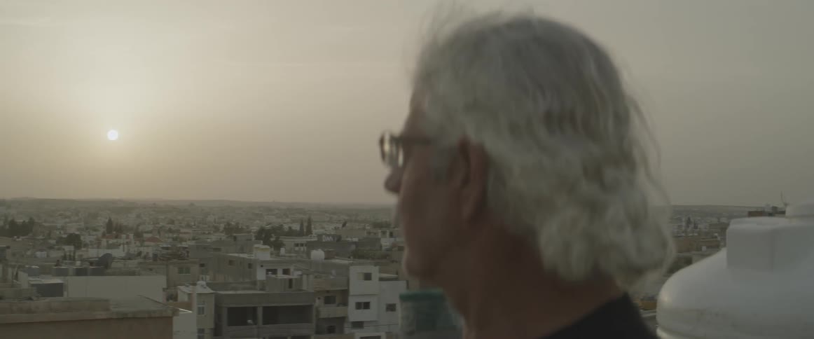 An older man with glasses staring out over a city from a balcony