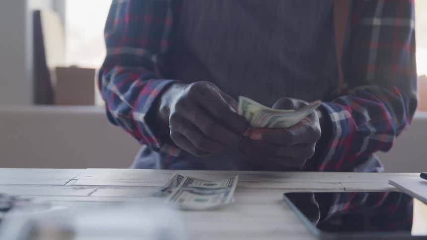 A man wearing a plaid shirt sits at a light colored table intently counting a stack of dollar bills with his hands while more cash lies on the table next to a tablet