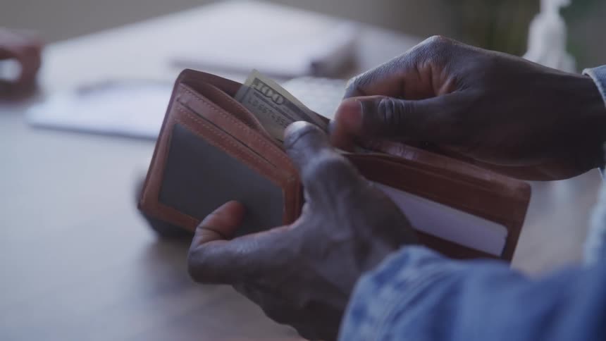 A pair of dark hands carefully opens a light brown leather wallet then pulls out a single united states dollar bill and hands it to another person whose hand comes into view to accept the money