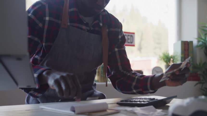 A man in a plaid shirt and apron sits at a desk counting money with a calculator and writing on a notepad with a sunlit window and plants in the background