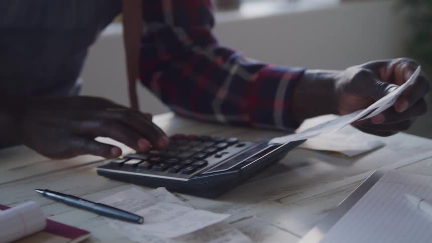 A person seen from the waist up wearing a plaid shirt sits at a wooden table using a calculator with one hand while holding receipts and papers with the other