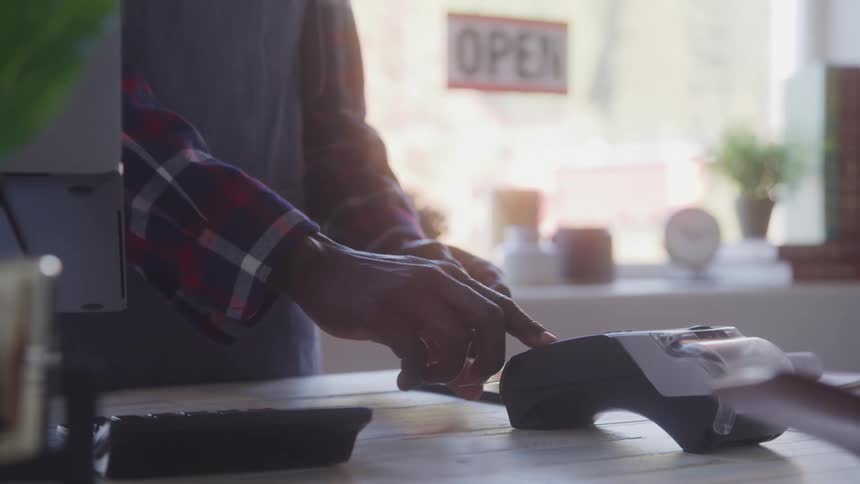 A barista in a plaid shirt hands a take away coffee cup to a customer then processes a credit card payment using a point of sale terminal on a counter with an open sign visible in the window
